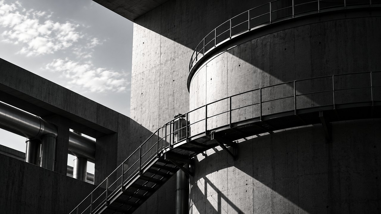 A concrete tank ringed by steel walkways, reached by an open steel stair, beside a concrete frame carrying large steel pipes.